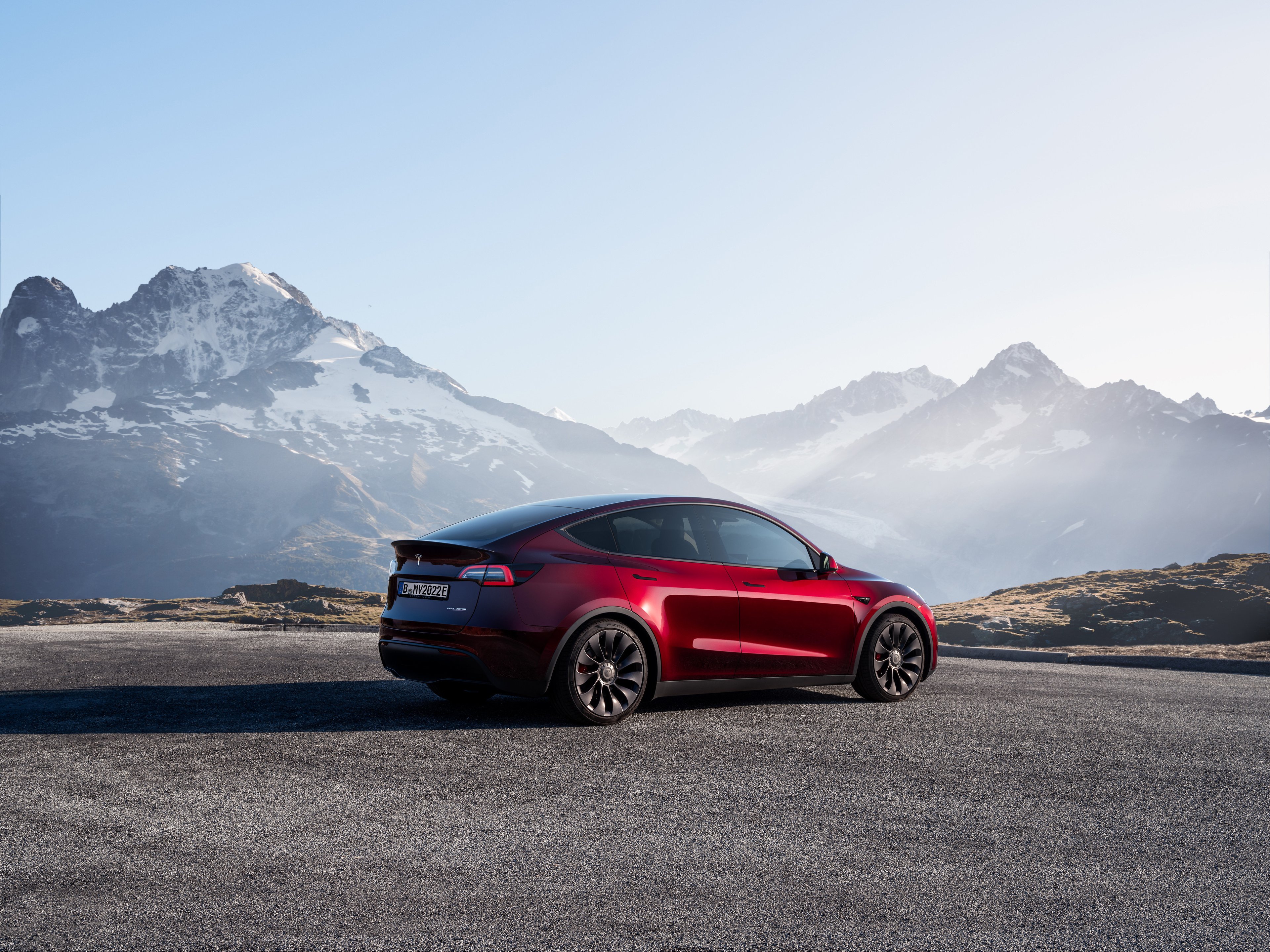 A red Tesla Model Y parked in a lot with a mountain range in the background.