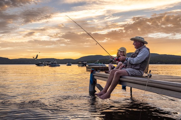 An adult and child sitting on a dock fishing at sunset.