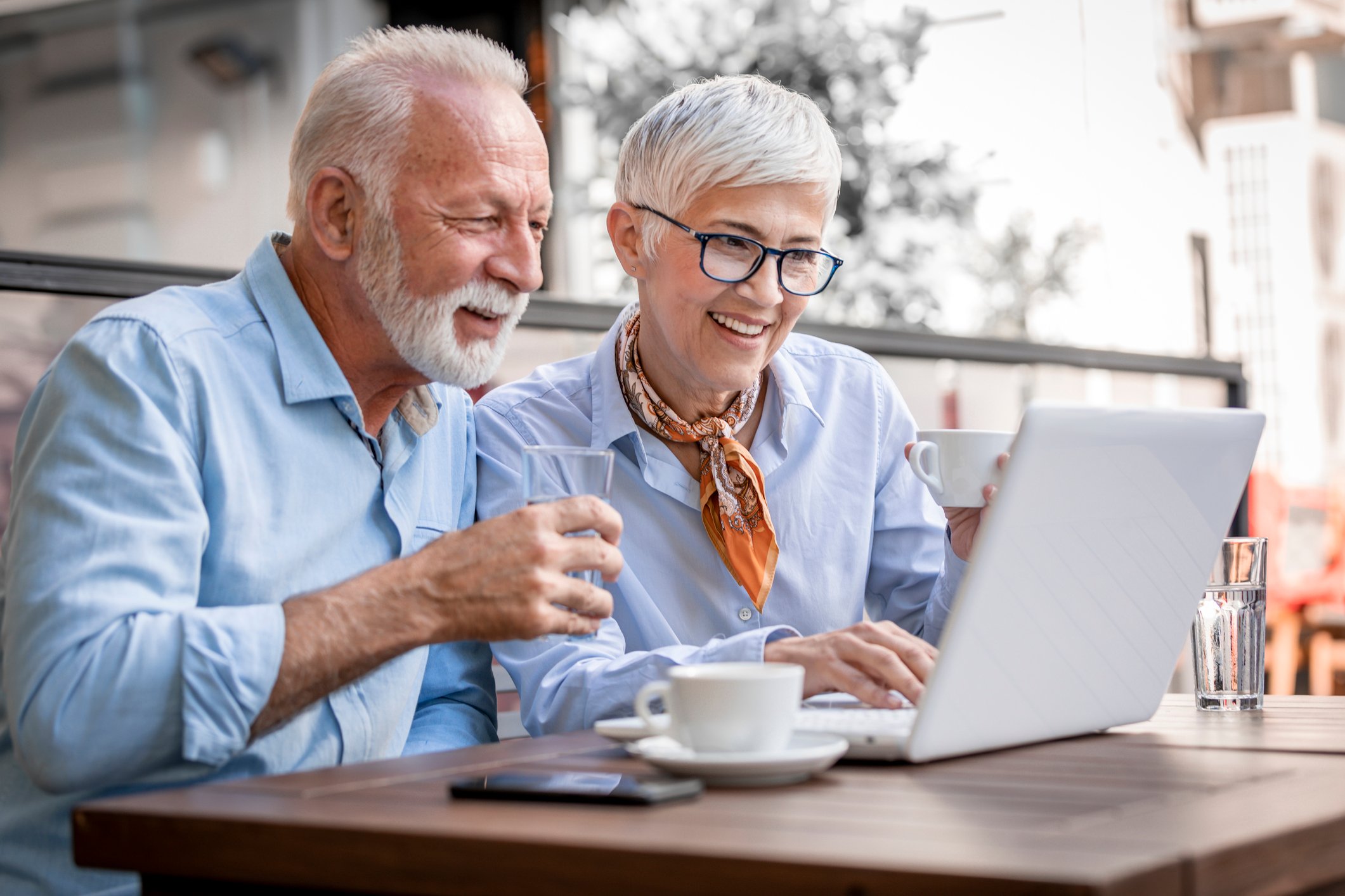 A retired couple smiles as they look at a computer screen while drinking coffee together.