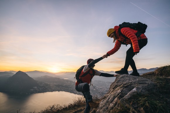 A hiker reaches out and helps another hiker up a mountain with a sunset in the background.
