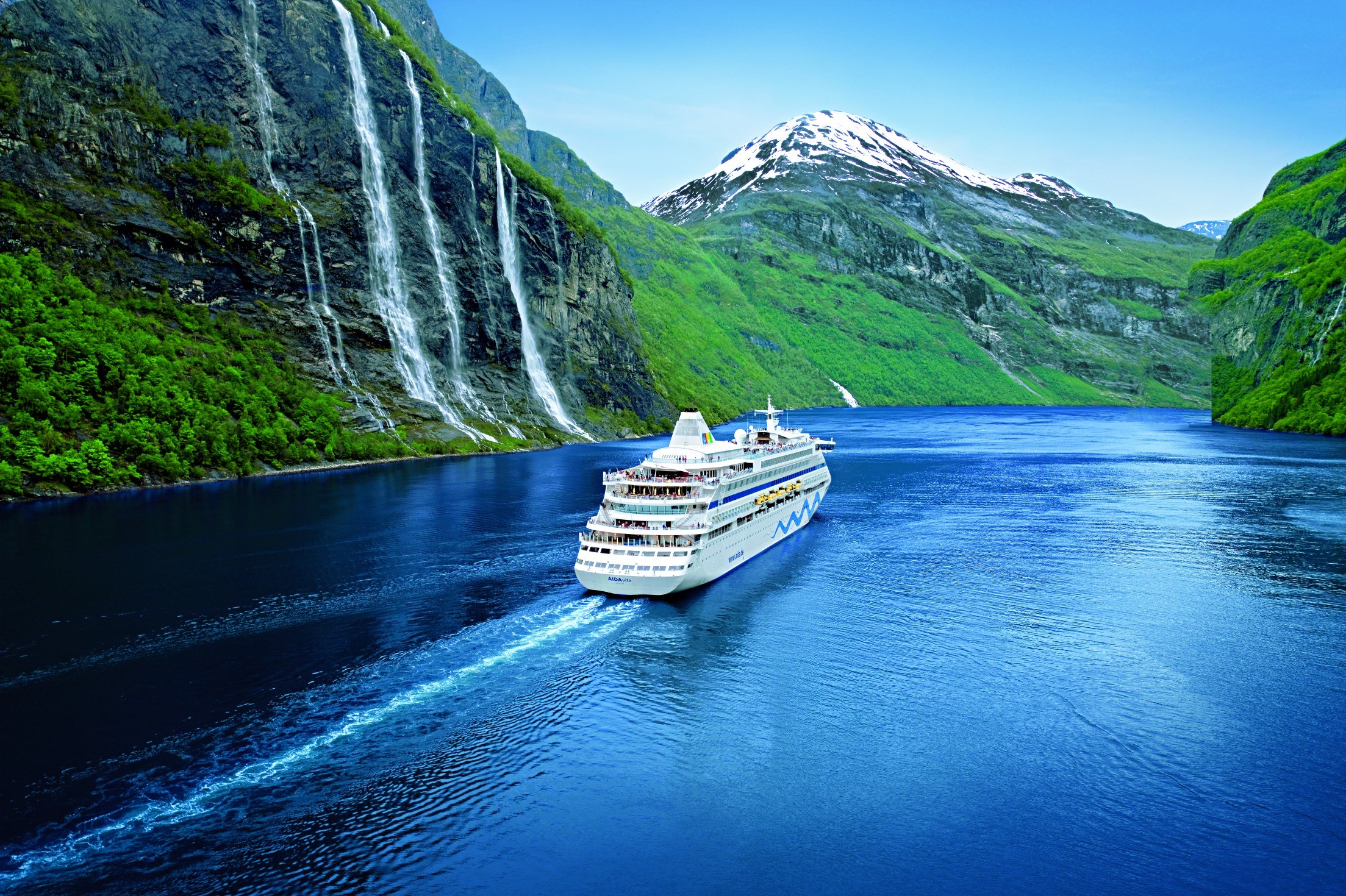 Cruise ship sailing through narrow passage with snowy mountain backdrop.