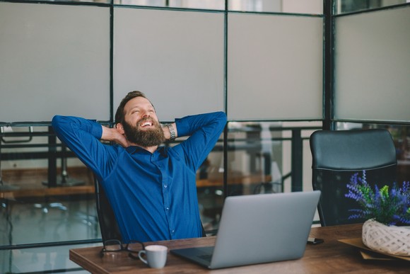Confident investor sitting at an office desk, reclining and smiling with their hands beind their head.