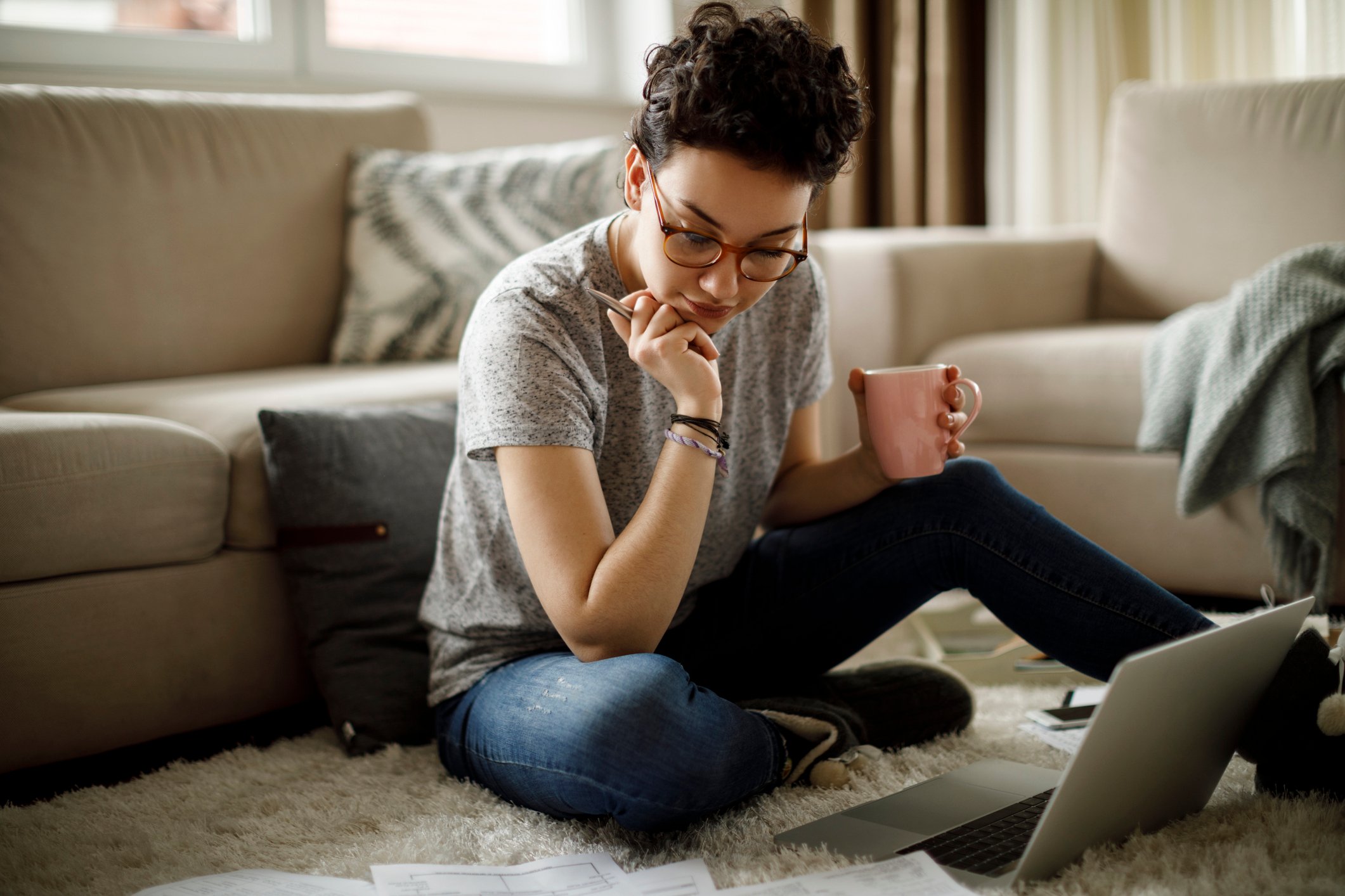 Serious person holding coffee mug and looking at documents spread out on floor.