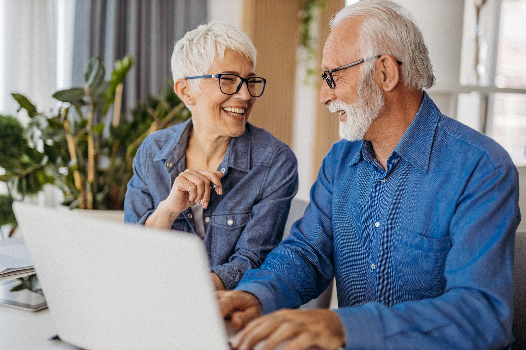 A retired couple smiling at each other while sitting at a table with a computer.