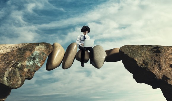 Person using a laptop computer sits on bridge between two cliffs, made of jammed-in rocks.