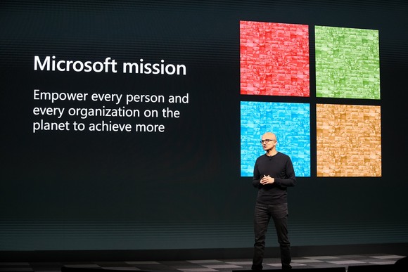Satya Nadella on stage in front of a screen with Microsoft's logo and mission.