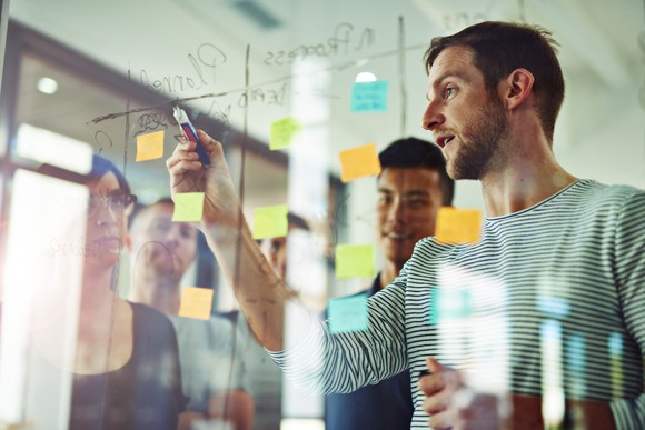 A group of people in an office setting watching a person write on a transparent dry-erase board.