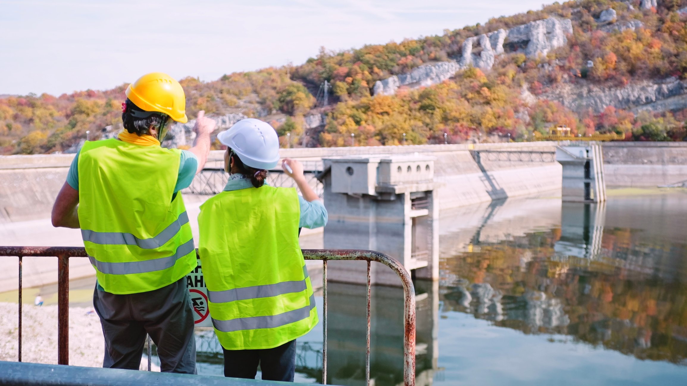 Two workers wearing hard hats and reflective vests survey a hydroelectric power station. 