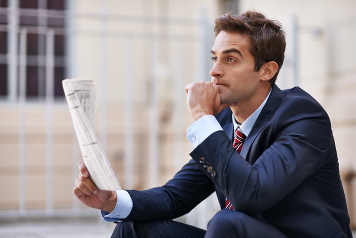 An person in a suit looks into the distance while holding a newspaper.