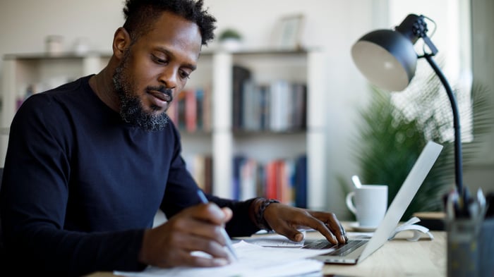 A person sits at a desk looking at papers on the desk and at a laptop computer. Also on the desk are a coffee cup and a desk lamp