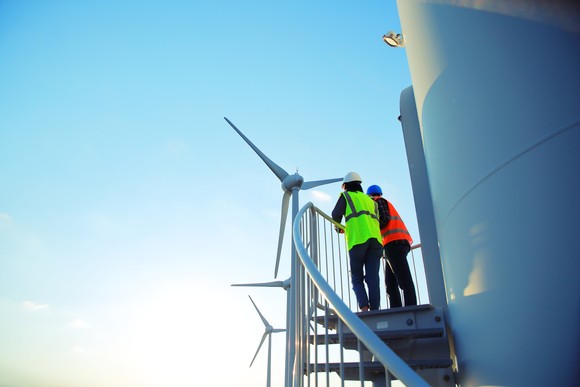 Two workers wearing reflective vests and hard hats climb a staircase next to a row of wind turbines. 