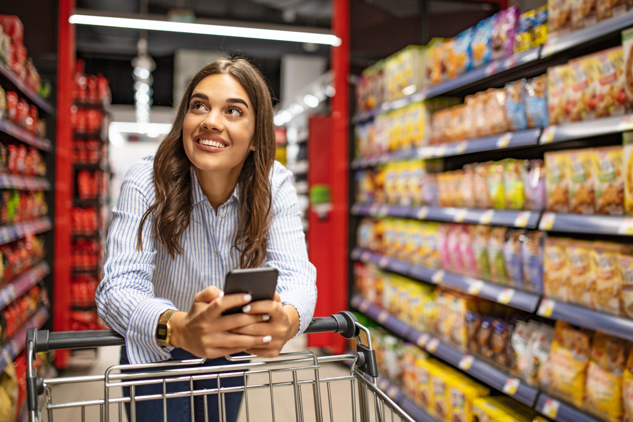 A shopper pushes a grocery cart through an aisle while holding a smartphone.