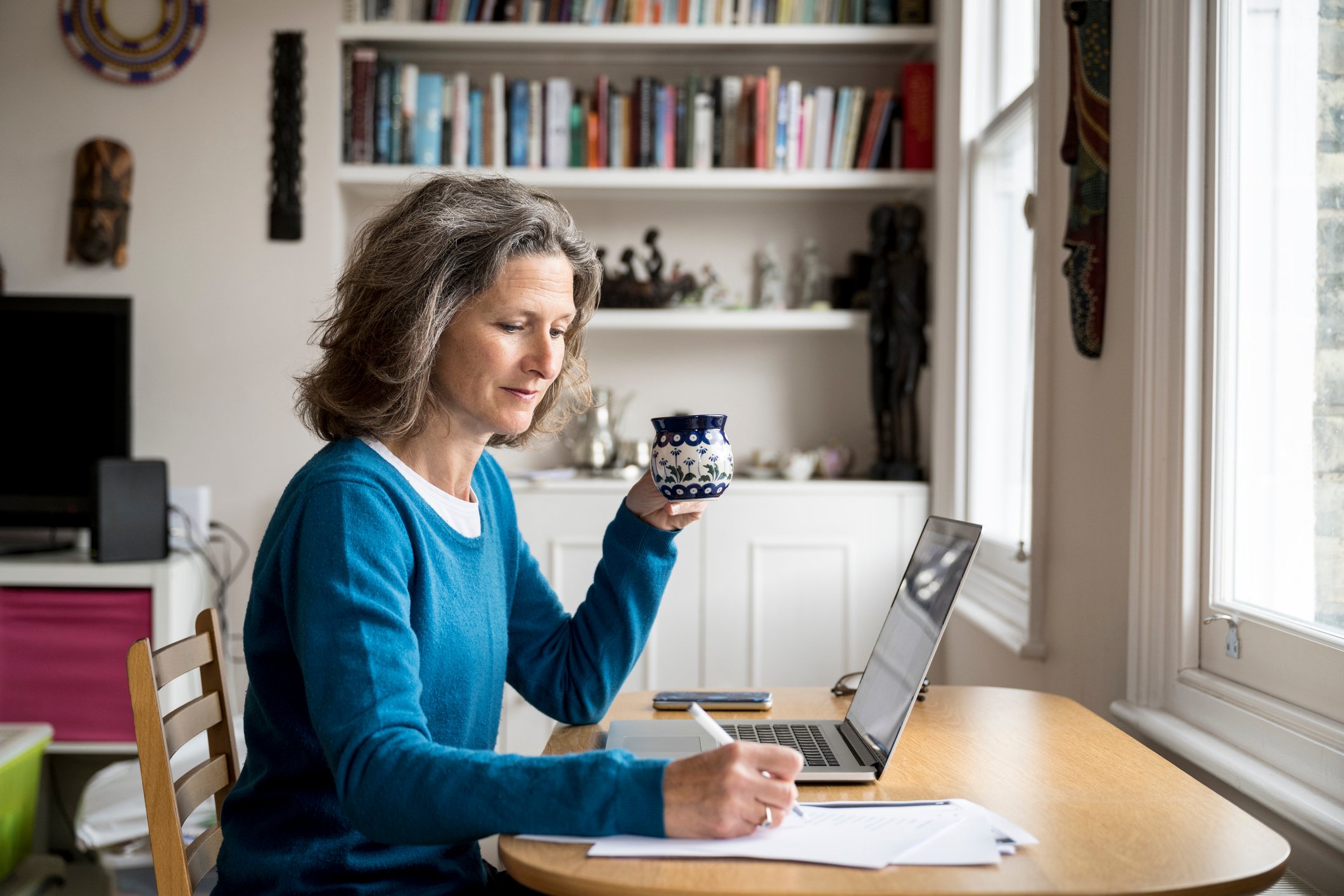 Someone sitting at a table writing on paper with a coffee cup in one hand.