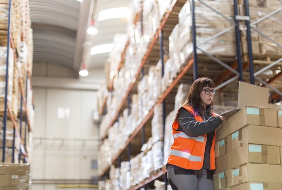 A warehouse worker on a laptop surrounded by packages. 