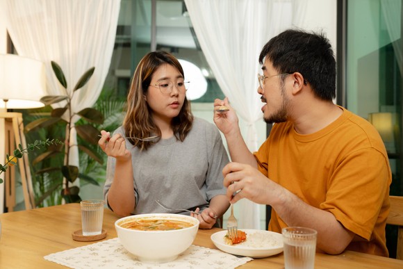Two people sit at a table in a room eating soup. Also on the table are a bowl and glasses