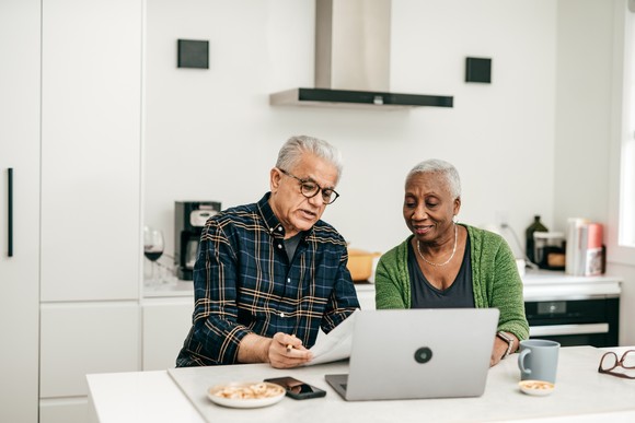 A retired couple sits at the kitchen table with a computer, where they are reviewing financial documents.