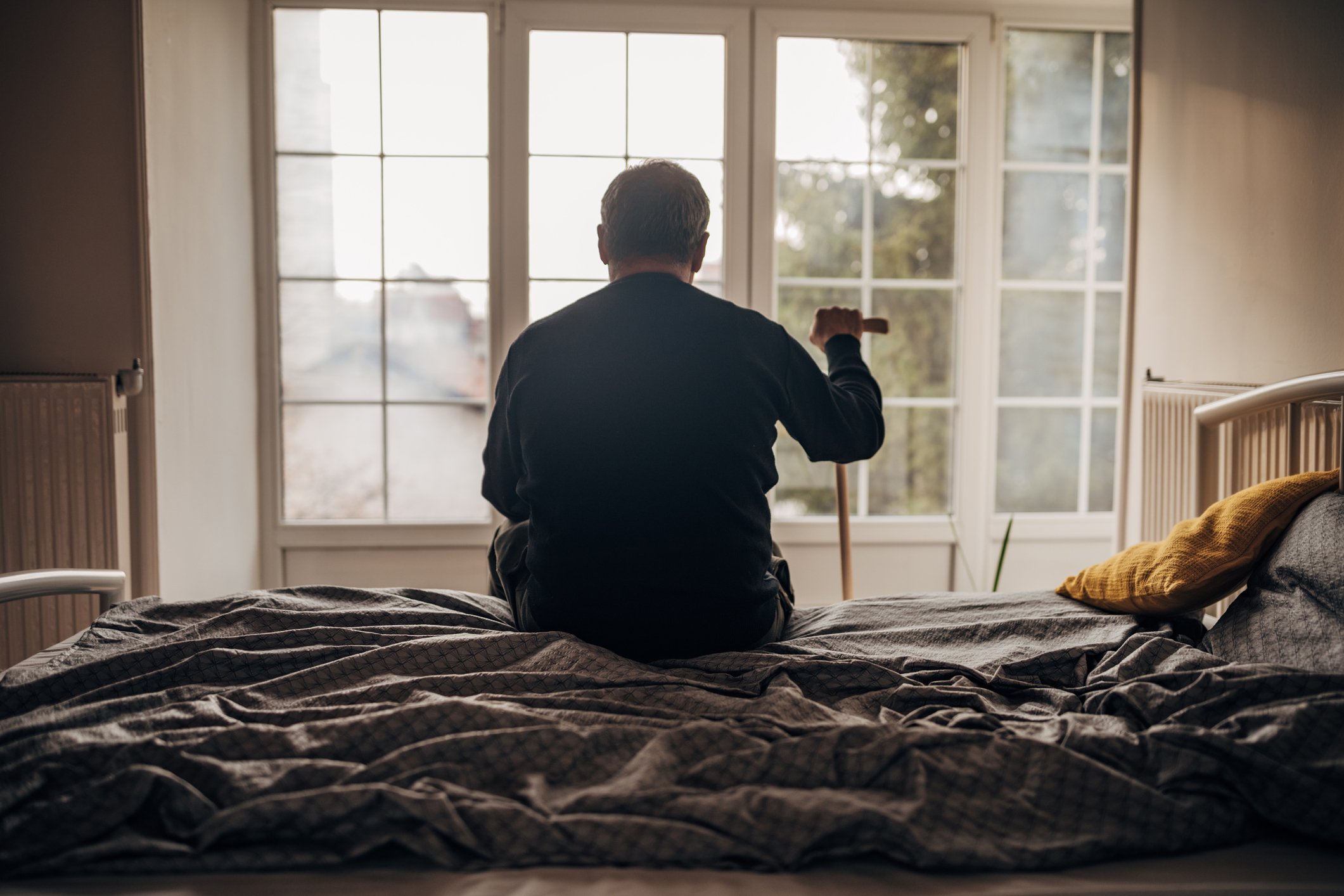 Person sitting on bed, holding cane, and looking out a window.