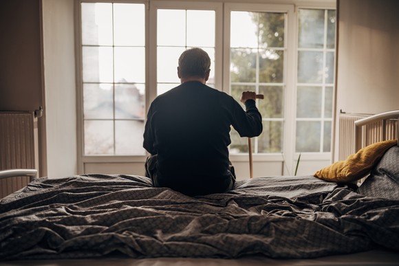 Person sitting on bed, holding cane, and looking out a window.