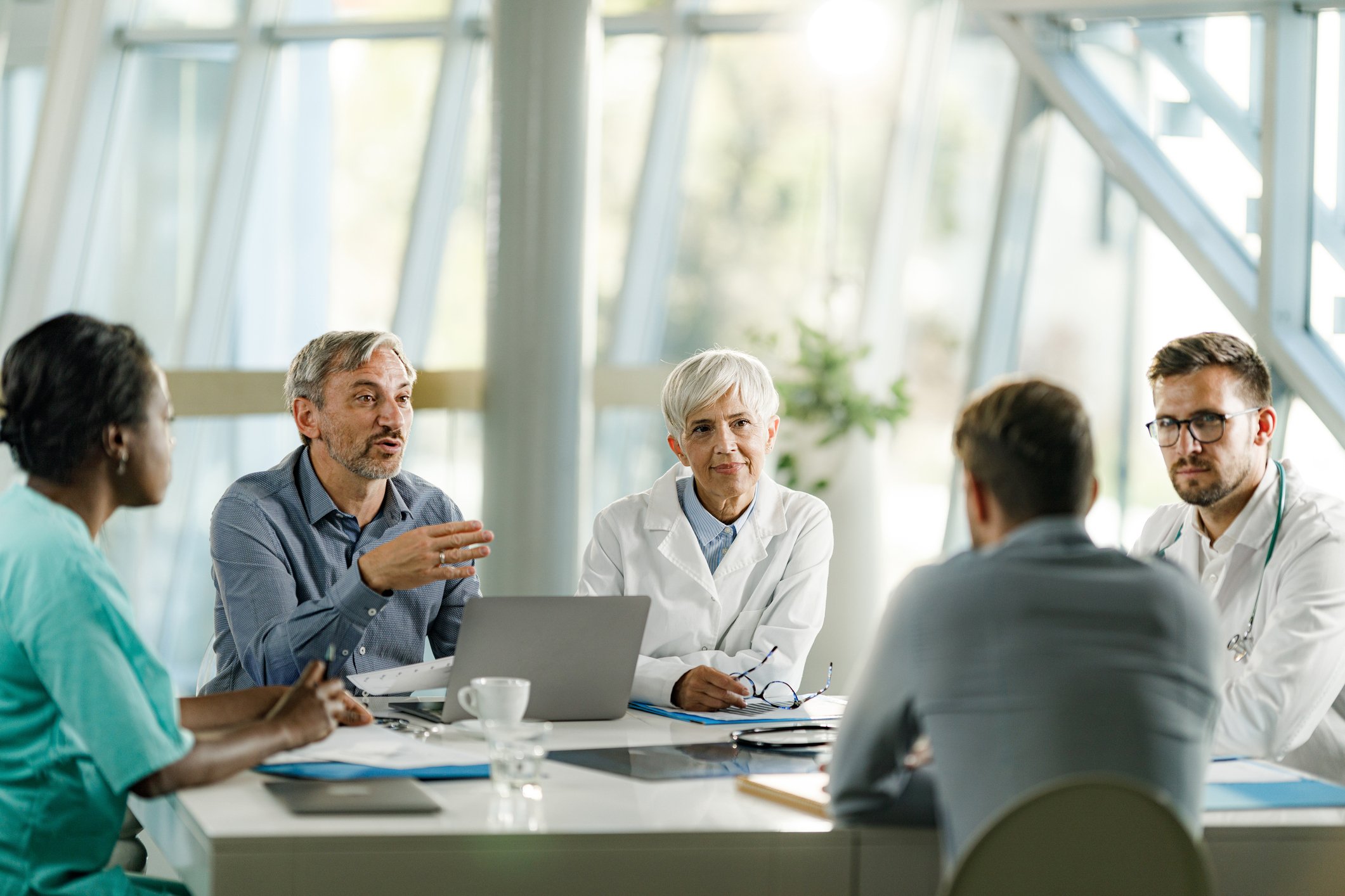 A group of doctors and a couple of their coworkers have a meeting at a table in an atrium.