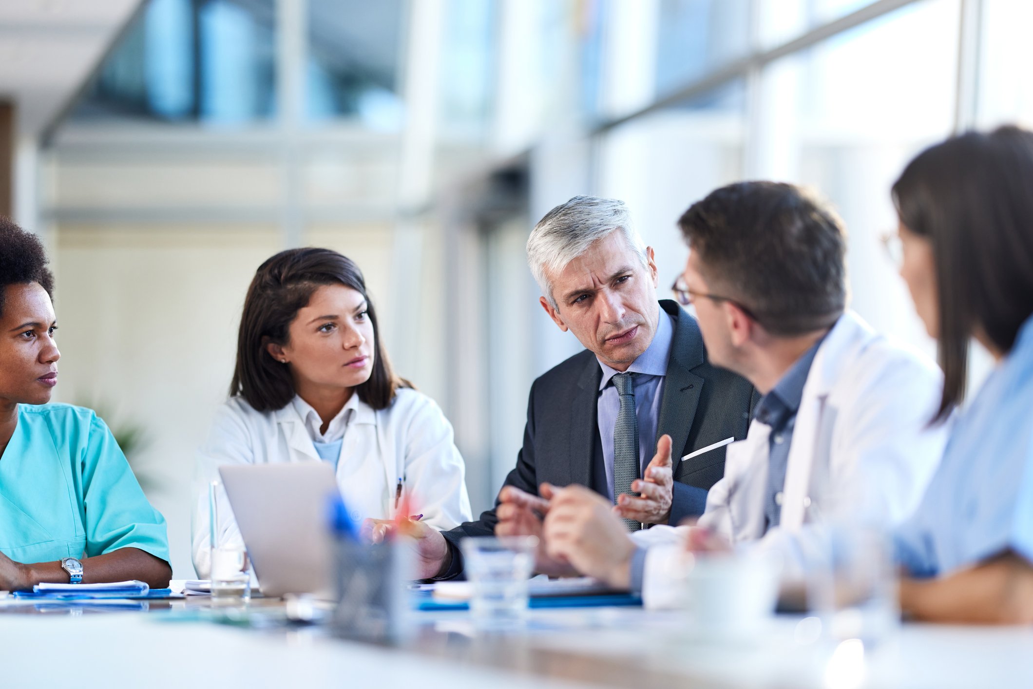An administrator talks with a team of doctors while sitting at a table in an atrium.