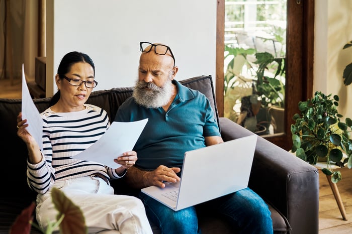 Two people on a couch, reviewing paperwork and using a laptop.