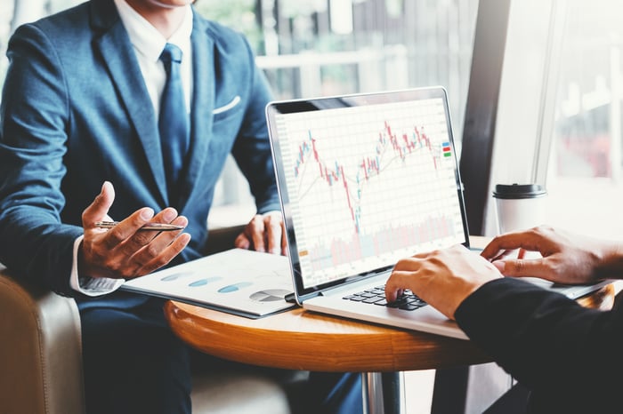 Two investors review financial data on a laptop while sitting at a table.