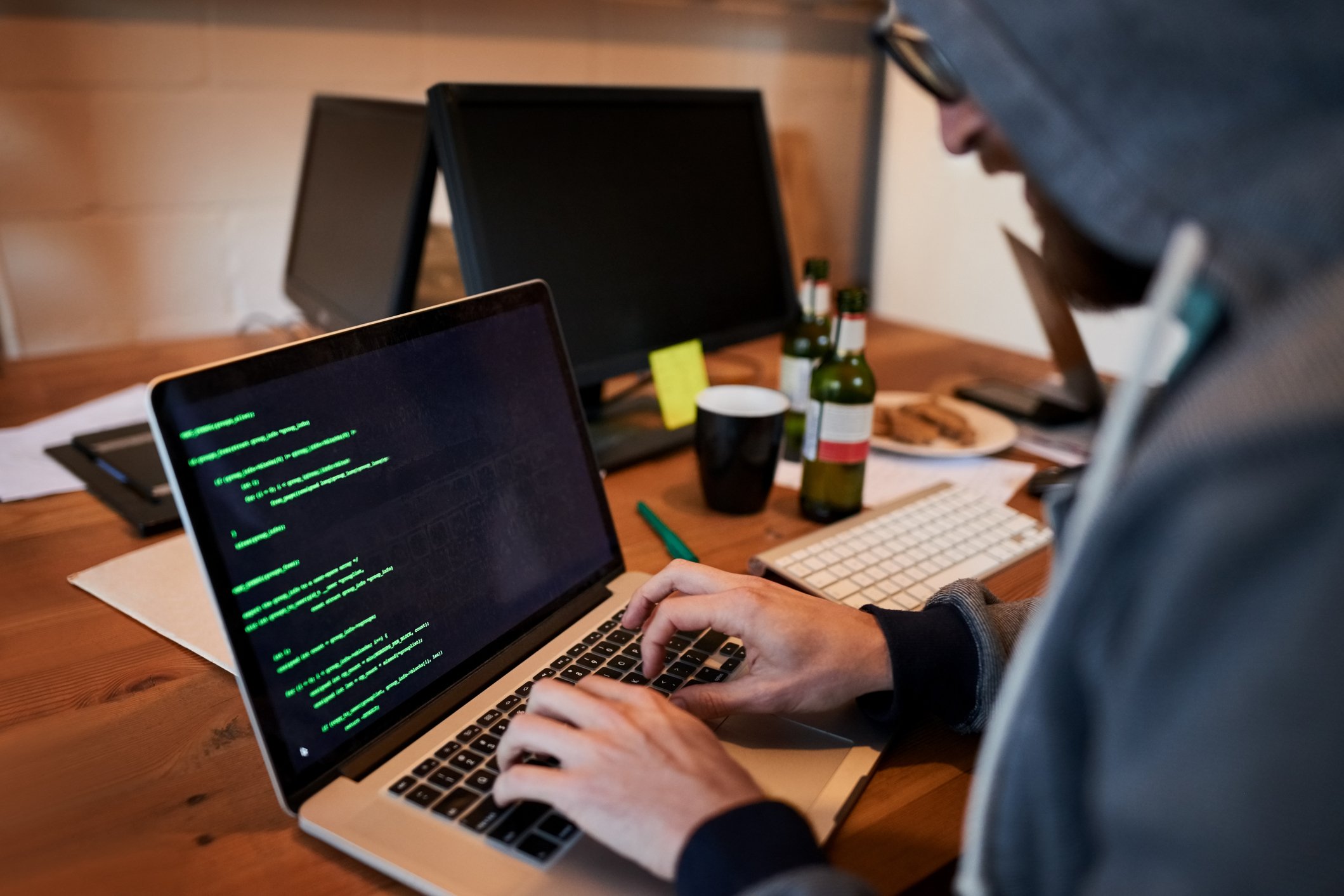 Hacker wearing a hooded sweatshirt while writing code on a laptop computer.