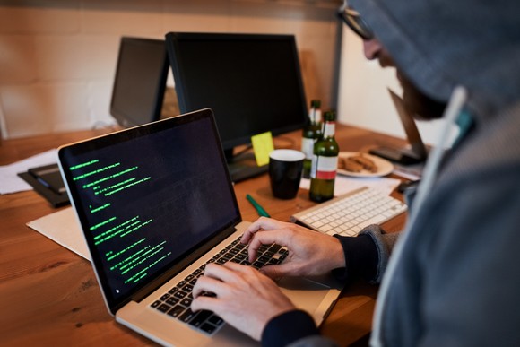 Hacker wearing a hooded sweatshirt while writing code on a laptop computer.