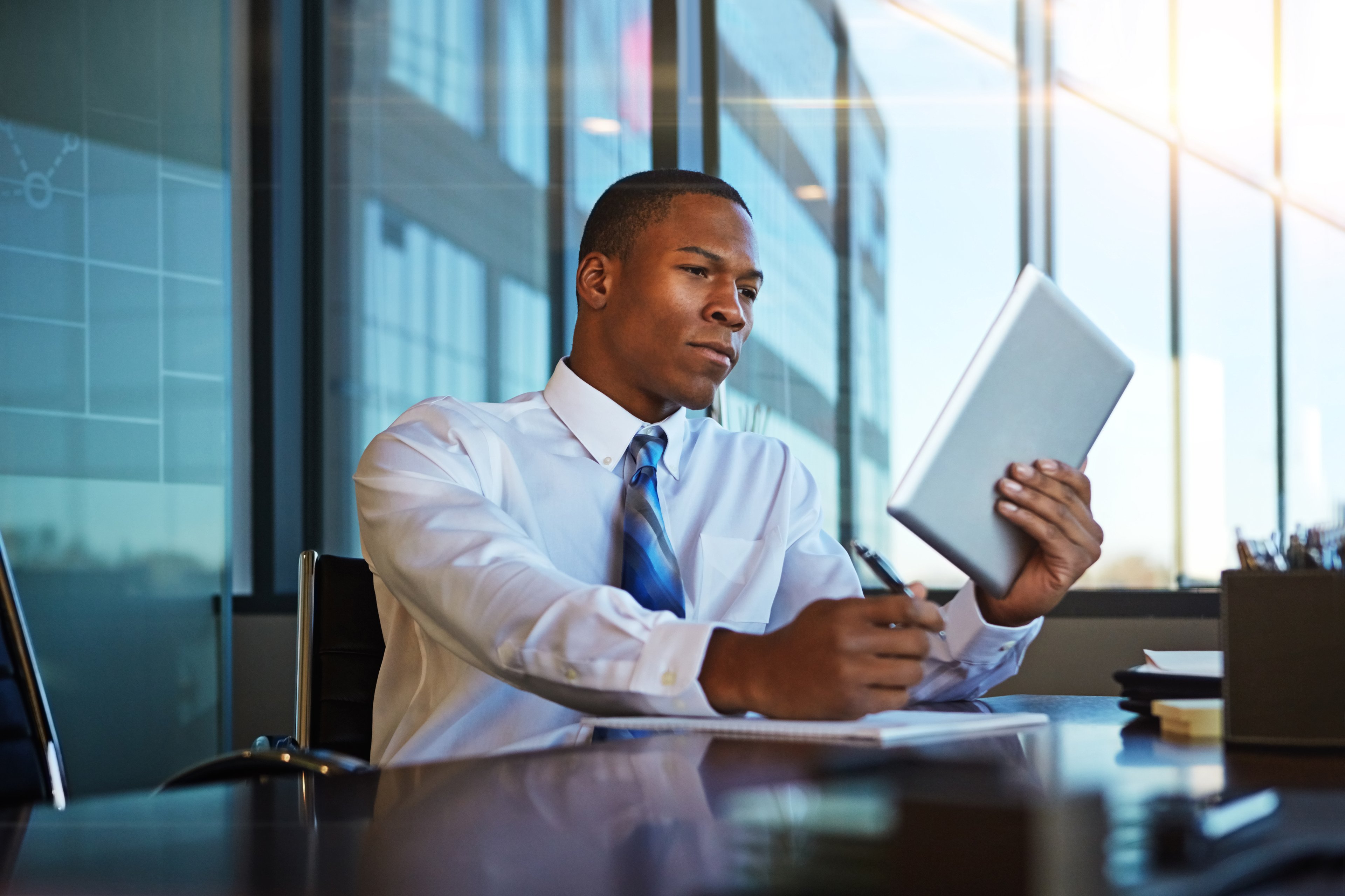 Male investor in dress clothes looks at a tablet and makes noted on a pad. 