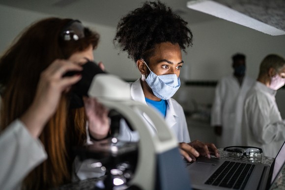 A pair of scientists work at a lab bench, with one looking into a microscope while another looks at a laptop.