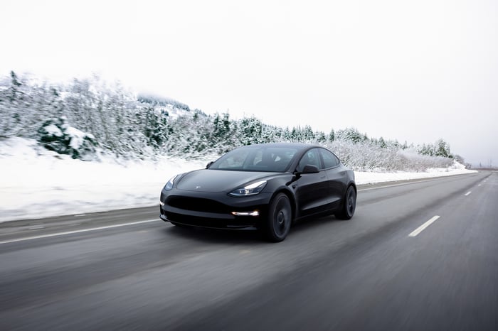 An all-electric Tesla Model 3 sedan driving down a road in wintry conditions. 