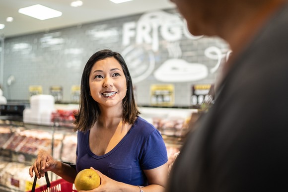 A person holding a piece of fruit and a shopping basket talking to someone in a supermarket.