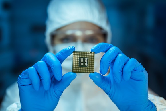 Semiconductor engineer in a clean suit and rubber gloves holding a microchip in front of their face.