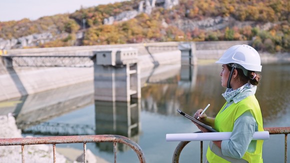 A person wearing personal protective equipment holds a clipboard while looking at a hydroelectric facility. 