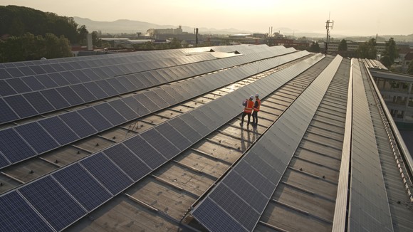 Two workers wearing personal protective equipment stand on top of a roof covered with solar panels. 