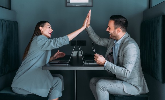 People in suits sitting at a table, high fiving over laptop computes.