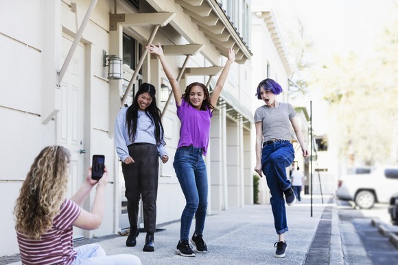 Person using smartphone to film three friends dancing on a sidewalk.