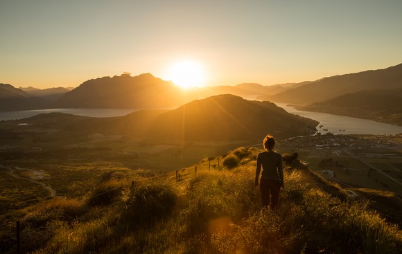 A person stands on a grassy mountain looking out over a mountain range at the rising sun. 