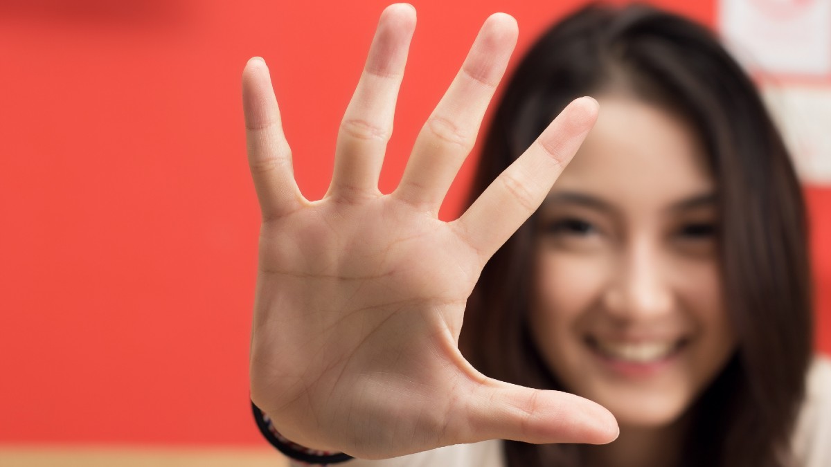 A hand is wide open, with five fingers out. Behind it is a blurry smiling face.