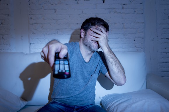 Person points a TV remote at an unseen screen, looking away and covering their face with the other hand.
