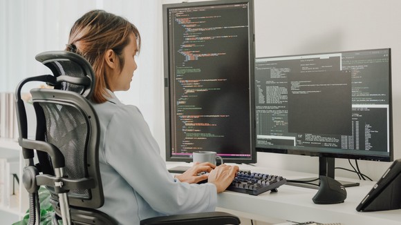 Software engeineer seated at a desk, writing code on two different computer monitors.