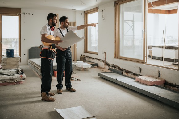Two construction workers discussing plans in a partially-built house.