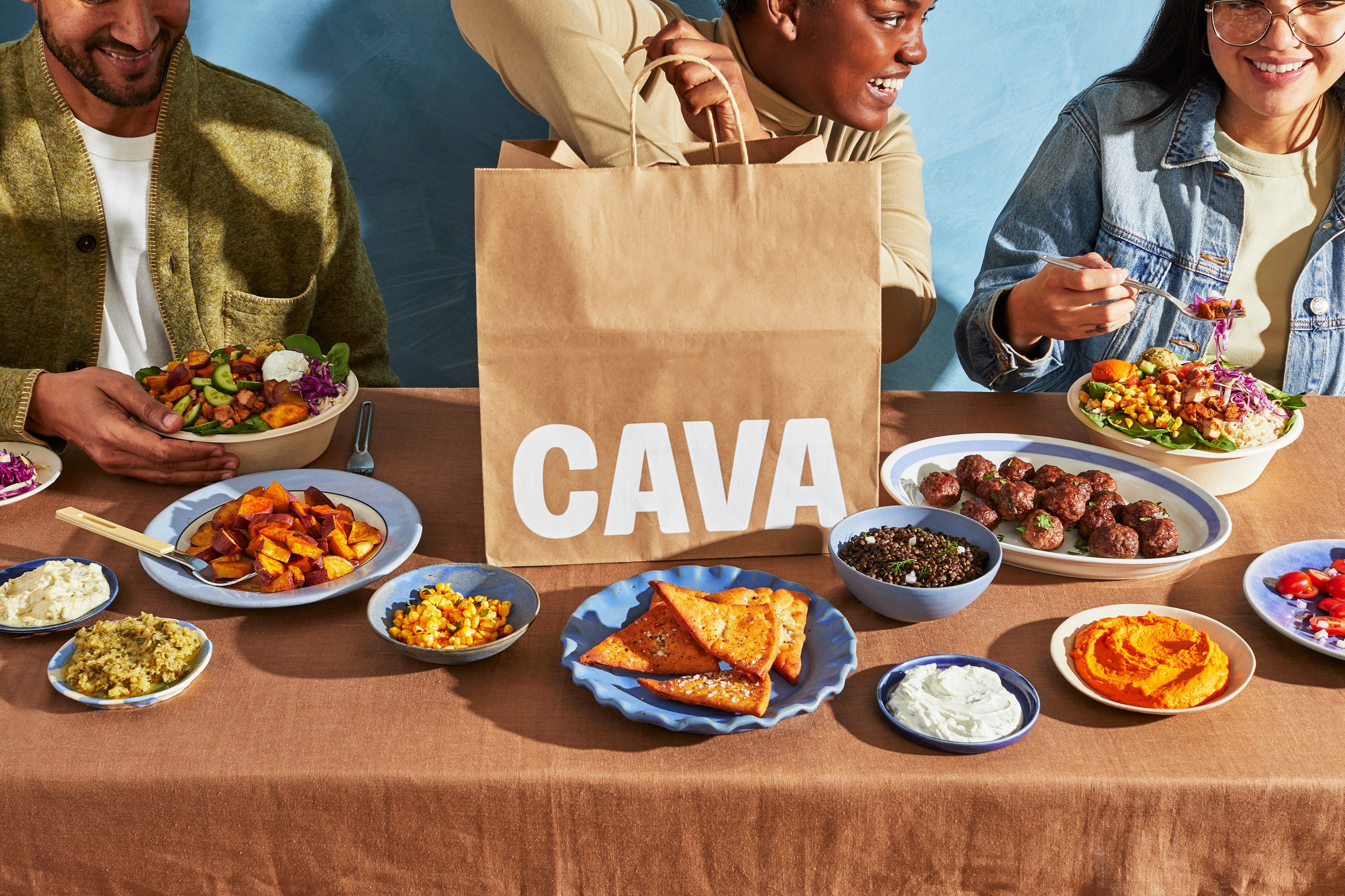 Several people sitting at a table full of food while one takes more food from a Cava Restaurant bag.