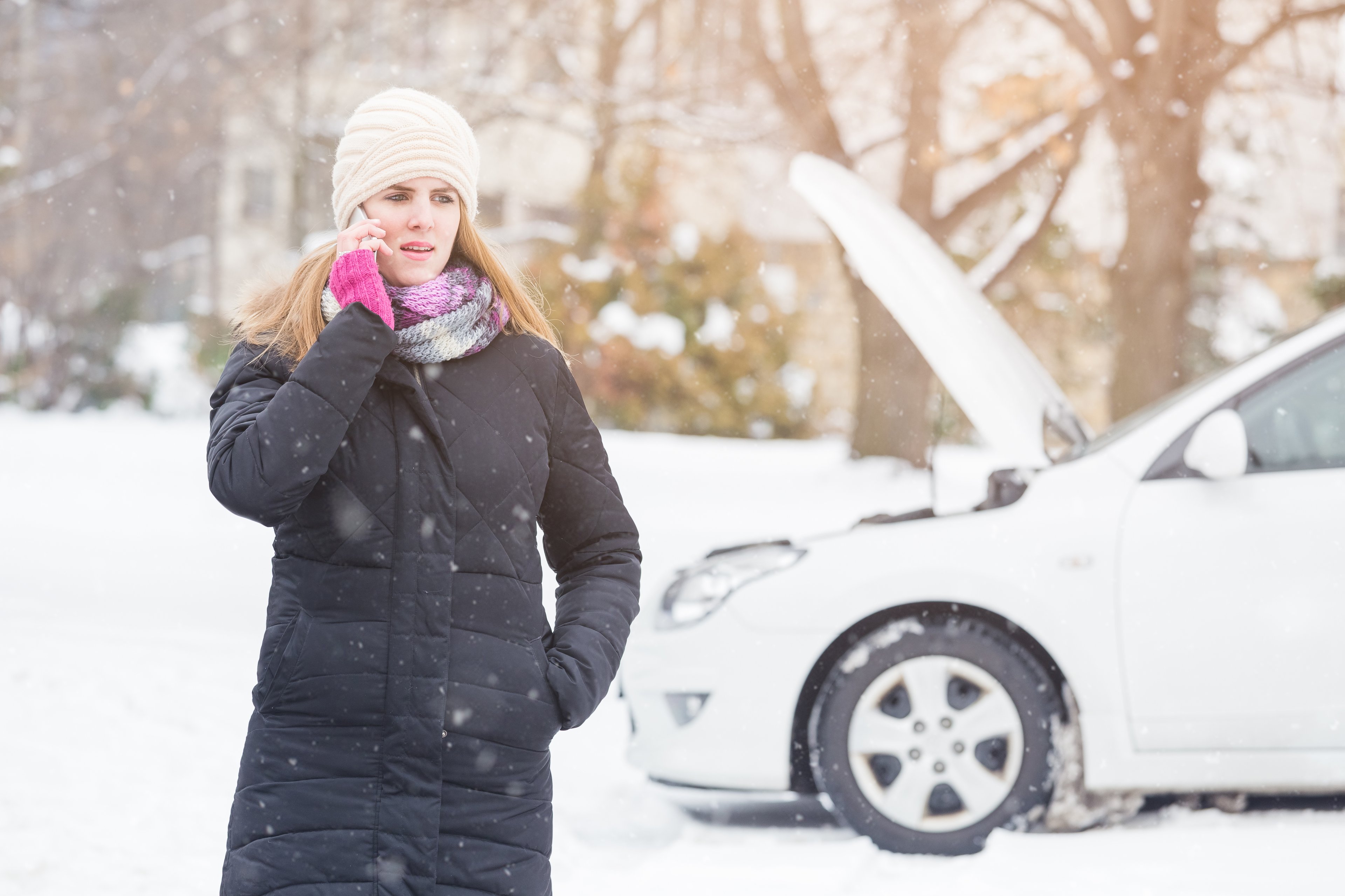 Person on phone in front of car with the hood open during winter storm.