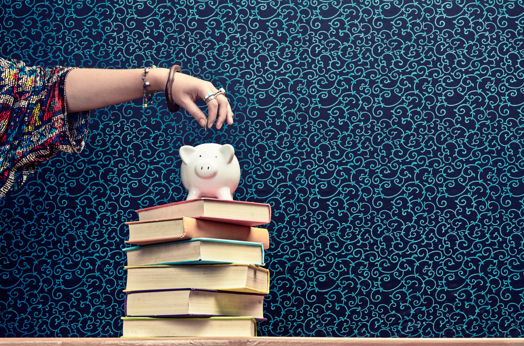 Hand putting coin in piggy bank atop a stack of books.