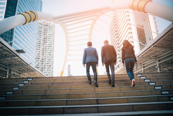 Three people walking up a staircase toward a bridge with sunlight pouring in. 