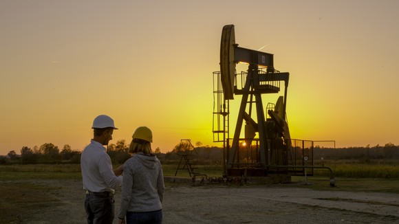 Two people wearing hard hats overlook a pumpjack at sunset. 