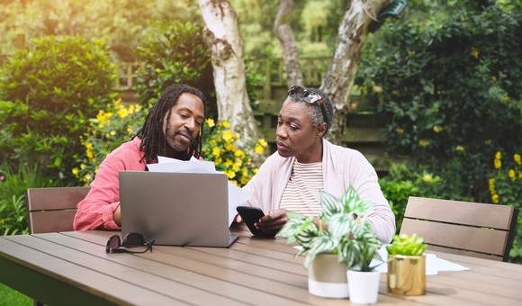 Two people seated at an outdoor table and looking at a laptop.