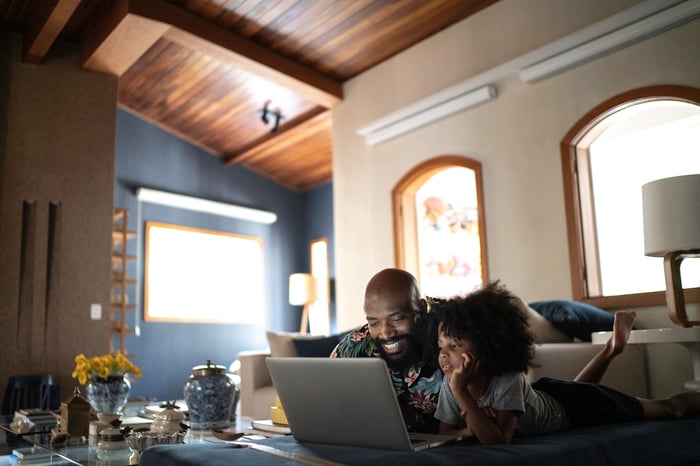 A man and a child watch content on a laptop on a coffee table in a living room.