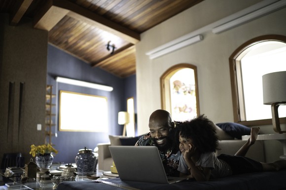 A man and a child watch content on a laptop on a coffee table in a living room.